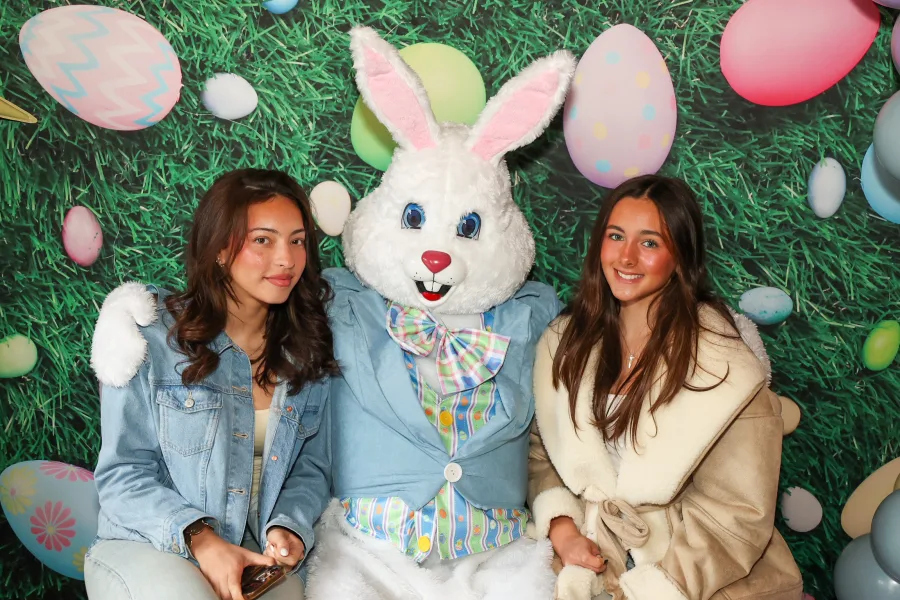 Two students posing with the Easter Bunny at a staged photo op backdrop