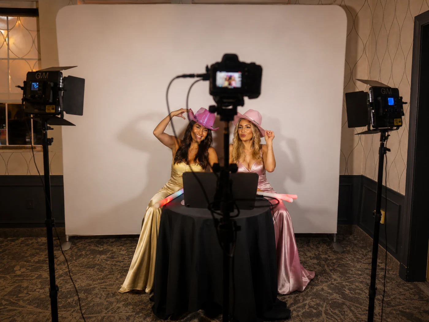 The Confessional Booth setup seen from behind the camera: clean backdrop, two GVM studio lights on stands, tap-to-start screen and tripod-mounted camera with two guests recording