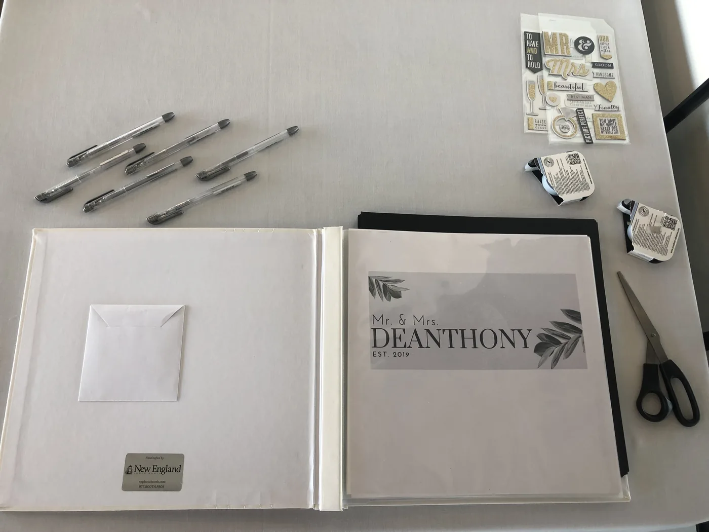 An open guest book on a table beside silver gel pens, scissors and sheets of embellishments